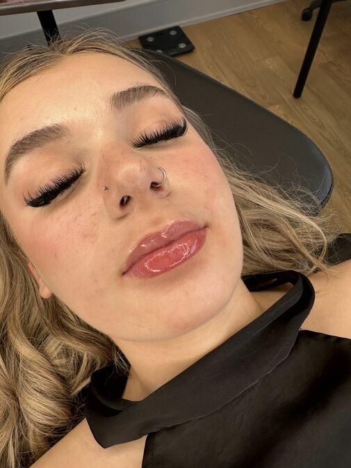 A person with long eyelashes, glossy lips, and a nose piercing is lying back on a chair, eyes closed, in a well-lit indoor setting—perfect for our Before and After Gallery Vancouver.