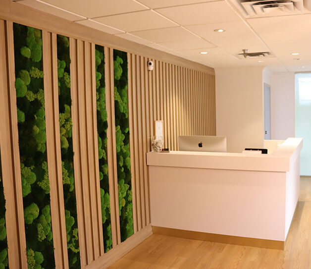Modern office reception area at Broadway Anti-Aging Clinic with a white desk, wood flooring, and a wall featuring vertical wooden slats and green plant accents. A computer and small decor items sit on the desk.