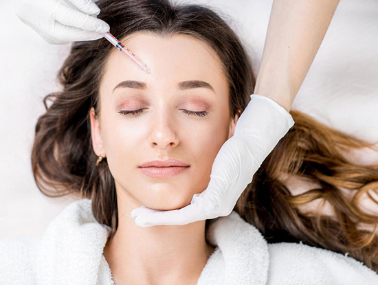 A person with closed eyes receives a Botox injection in the forehead from a gloved professional, while another gloved hand gently supports their chin.