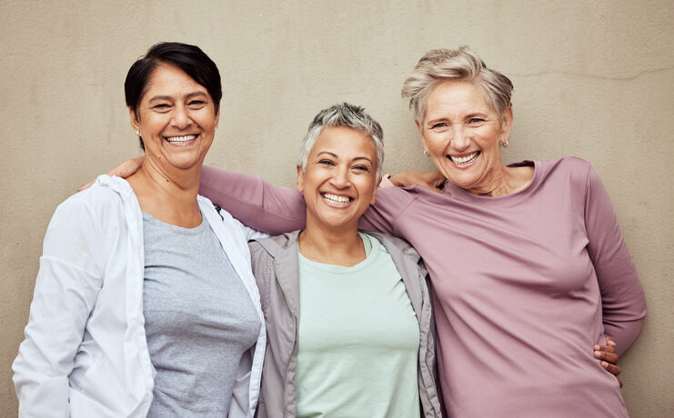 Three older women stand together, smiling at the camera with one woman’s arm around the others, radiating joy and confidence—perhaps a testament to successful hormone optimization—against a plain beige wall.