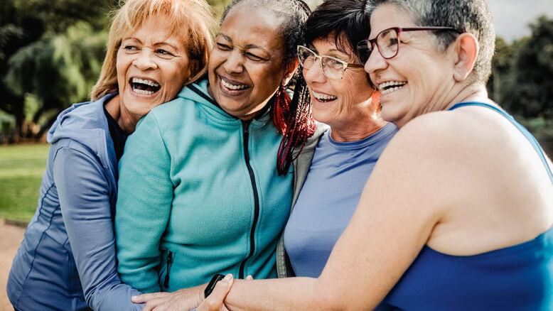 Four older women stand outdoors closely together, smiling and hugging each other in casual athletic wear, celebrating the benefits of hormone optimization and healthy living.
