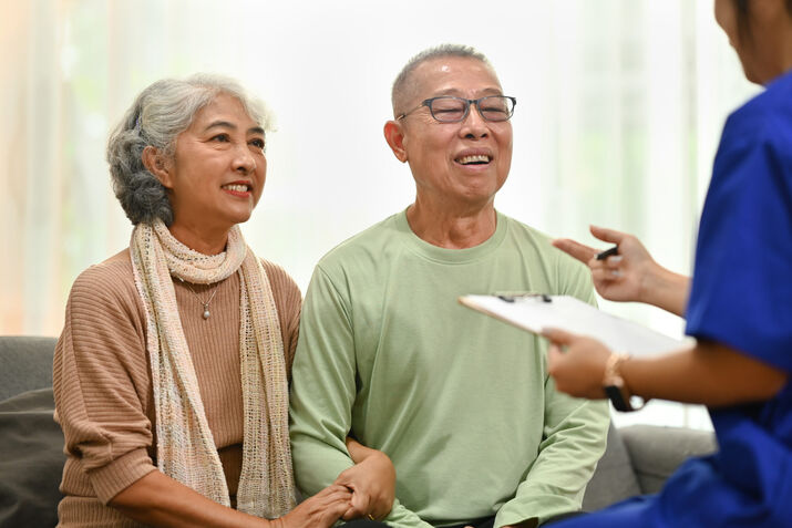 An older couple sits on a couch, smiling and holding hands, while a healthcare professional in blue scrubs discusses hormone optimization and takes notes on a clipboard.