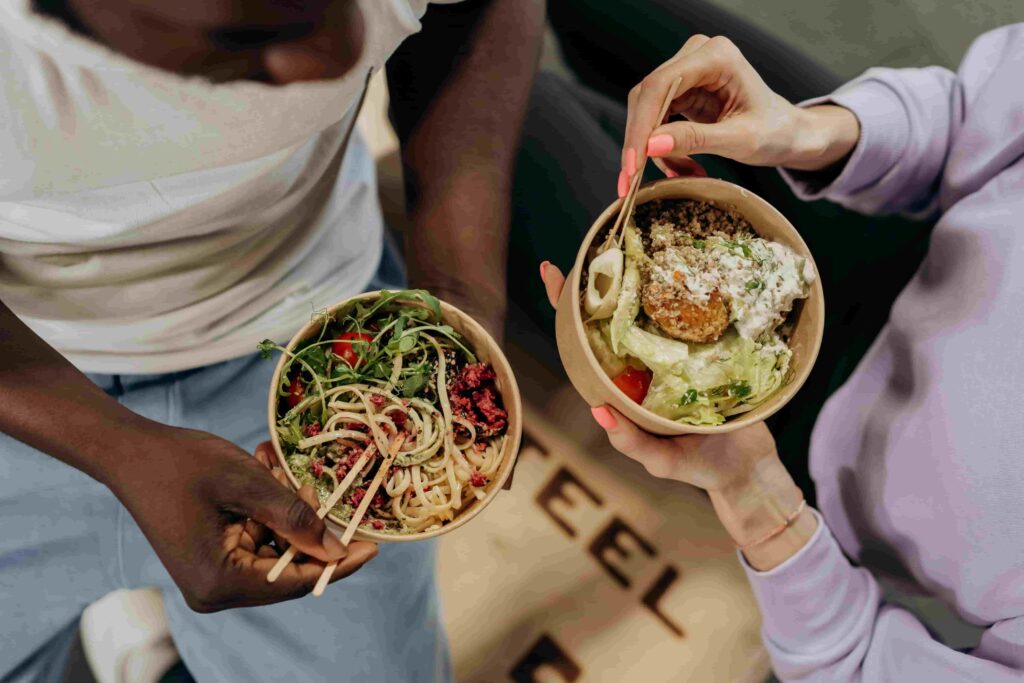 Two people enjoying bowls of fresh food inspired by the Longevity Diet—one with noodles and greens, the other with salad and a breaded item—eating together with chopsticks and a fork.