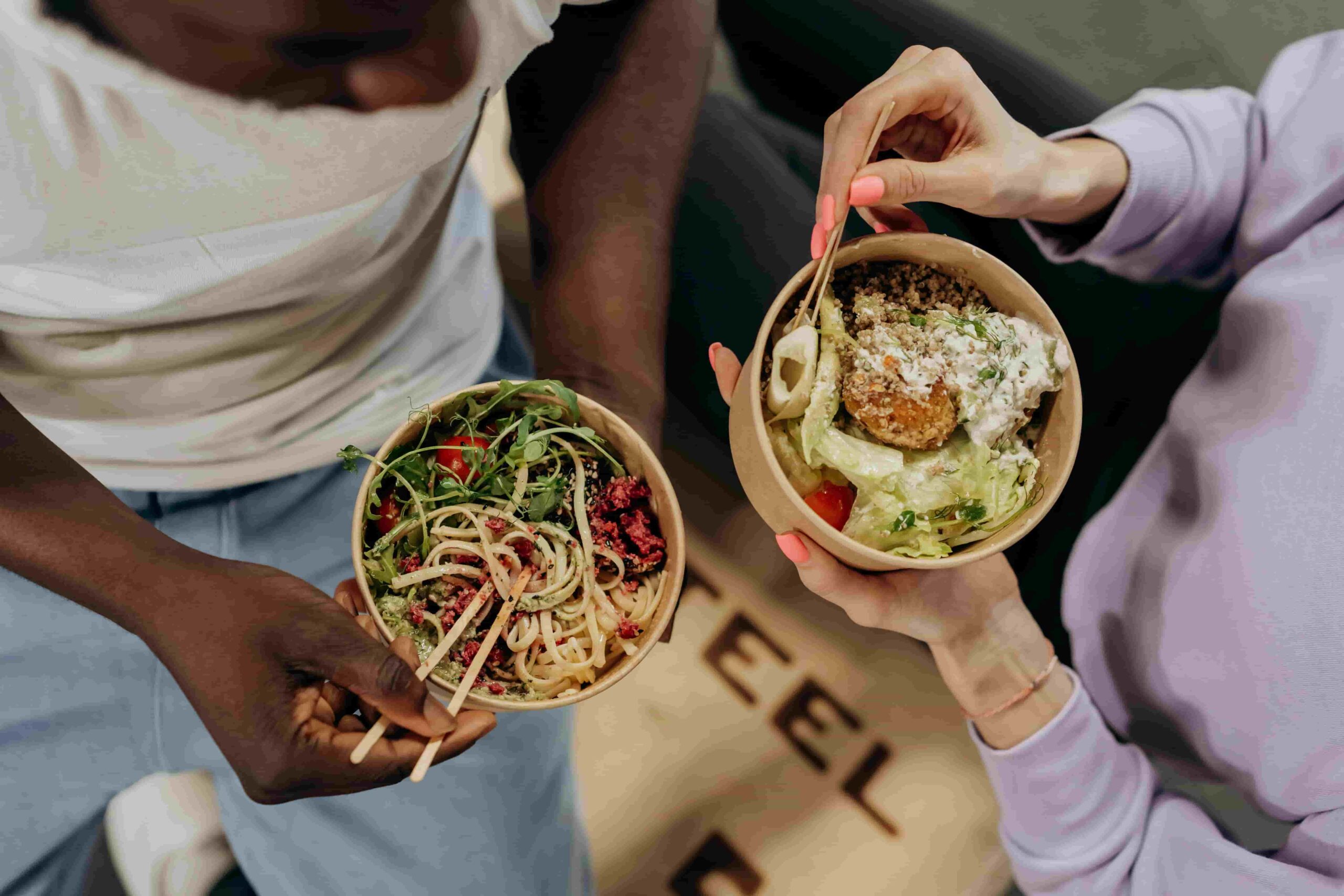 Two people enjoying bowls of fresh food inspired by the Longevity Diet—one with noodles and greens, the other with salad and a breaded item—eating together with chopsticks and a fork.