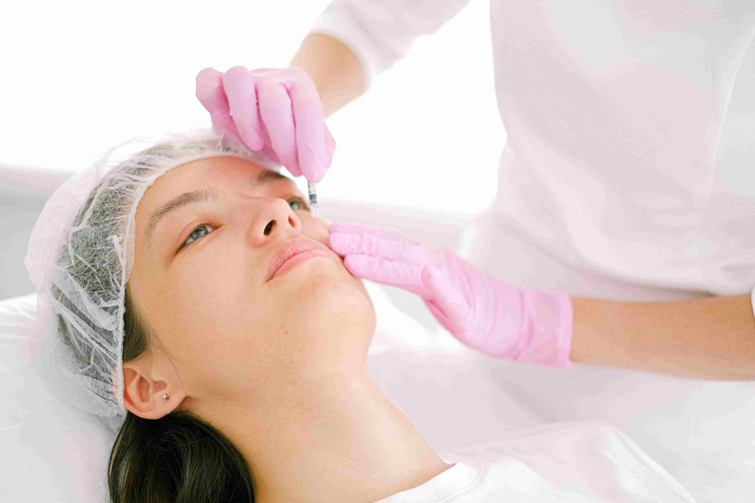A person wearing a hair cap receives a facial treatment from a professional in pink gloves, similar to those seeking natural-looking results—like asking, "Who is the best Doctor in Vancouver for natural-looking Masseter Botox?.