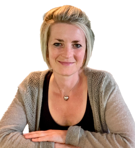 A woman with short blonde hair, wearing a gray cardigan over a black top and a heart-shaped necklace, sits at a table and smiles at the camera, radiating the confidence and vitality often seen in clients of Broadway Anti-Aging Clinic.